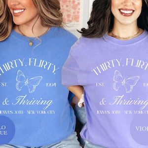 May include: Two t-shirts with a butterfly graphic and the text "Thirty, flirty & thriving" in white. The shirt on the left is blue and the shirt on the right is purple. The text "Flo Blue" is in a circle on the left shirt and the text "Violet" is in a circle on the right shirt.