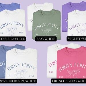 May include: Six folded t-shirts in different colors with the text "THIRTY, FLIRTY EST 1994" and a martini glass graphic. The colors are: Flo Blue/White, Bay/White, Violet/White, Washed Denim/White, and Crunchberry/White.