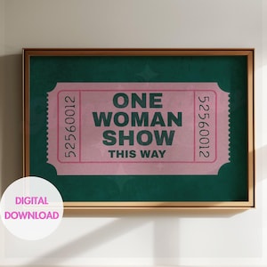 May include: A framed print featuring a pink ticket design with the words "ONE WOMAN SHOW THIS WAY" in green. The ticket has a serrated edge and the numbers 52560012 on each side. The background is a dark green color.