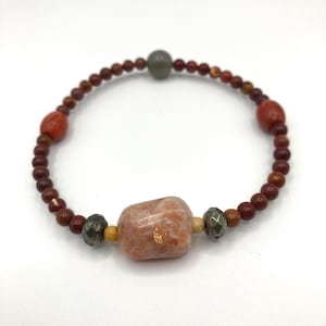 Könnte beinhalten: Ein Armband mit einer großen, rechteckigen, pfirsichfarbenen Stein mit Goldflocken. Das Armband enthält kleine, runde, rotbraune Perlen und größere, ovale, rote Perlen. Eine grau-grüne Perle ist oben zentriert.