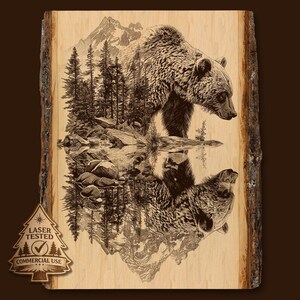 May include: A laser-etched wooden art piece featuring a grizzly bear standing near a lake, with its reflection visible. The design includes a mountain range, trees, and rocks. The artwork is on a wood slab with bark edges. The text "LASER TESTED" and "COMMERCIAL USE" is visible.