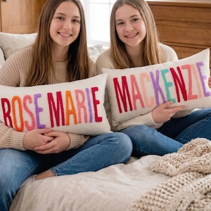 Two young women sitting on a bed, each holding a large pillow with the name "ROSE MARIE" embroidered on it.