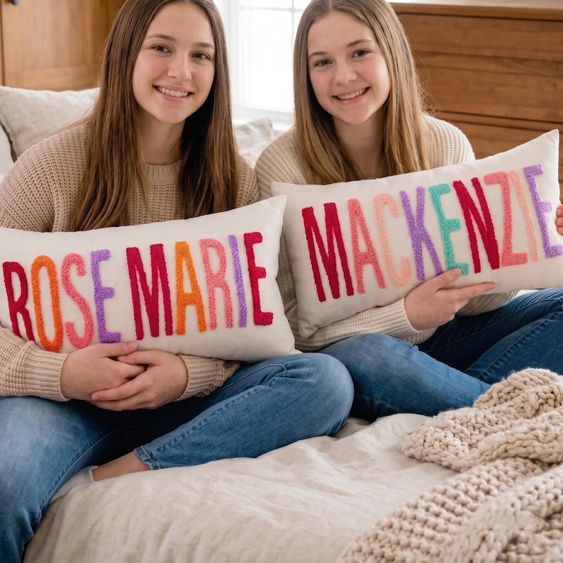 Two young women sitting on a bed, each holding a large pillow with the name "ROSE MARIE" embroidered on it.