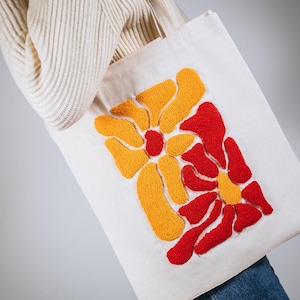 May include: A white canvas tote bag with a red and yellow embroidered floral design. The flowers are stylized and abstract.