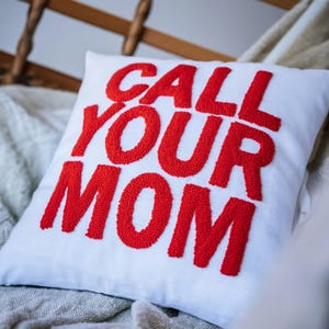 May include: White square pillow with the text "CALL YOUR MOM" in large, red, textured letters. The pillow is resting on a light-colored surface, with a wooden chair visible in the background.