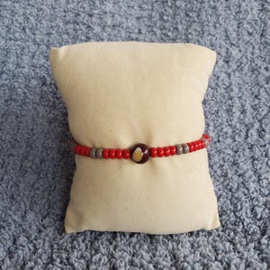Peut inclure: Un bracelet de perles composé d'un motif répétitif de petites perles sphériques rouges et grises. Le centre du bracelet est mis en valeur par une perle circulaire unique, plus grande, de couleur marron foncé avec un motif de bonbon maïs.