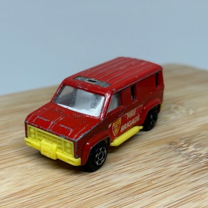 May include: Red toy fire truck van with a yellow bumper and yellow trim. The van has a white windshield and a black roof. The side of the van says "Fire Brigade" in white letters.