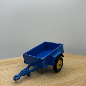 May include: A small, blue toy trailer with a rectangular bed and a single wheel. The trailer has a hitch and is painted in a solid blue color. The wheel has a yellow center and a black tire.