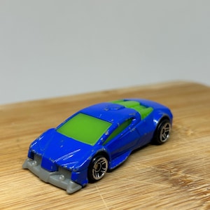 Pode incluir: Um carro de brinquedo azul com um pára-brisas verde e detalhes cinzentos. O carro tem rodas pretas e é feito de plástico.