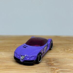 Può includere: Un'auto giocattolo viola con un parabrezza trasparente e ruote nere. L'auto ha il numero "24" sul lato e le parole "ALFA ROMEO" stampate sul lato.