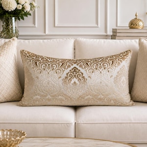 Può includere: Un cuscino lombare rettangolare con base color crema e un motivo damascato in velluto dorato in rilievo. Il design presenta volute ornamentali simmetriche e motivi floreali sulla parte anteriore, posizionato su un divano chiaro.