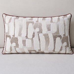 Puede incluir: Un cojín rectangular con un patrón abstracto y texturizado de formas blancas en relieve sobre un fondo beige y marrón claro. El cojín está acabado con un borde ribeteado en marrón oscuro que enmarca todo el diseño rectangular.