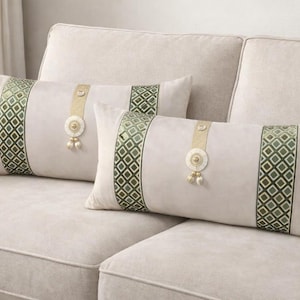 Peut inclure: Deux coussins décoratifs rectangulaires sur un canapé beige. Les coussins ont un fond beige clair avec un motif géométrique vert vertical à chaque extrémité. Une bande dorée et un médaillon décoratif avec des glands sont centrés sur chaque coussin.