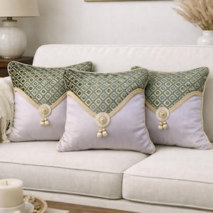 Peut inclure: Trois coussins décoratifs sur un canapé blanc. Les coussins ont une base violet clair avec une section supérieure à motifs géométriques verts. Des bordures et des glands dorés ajoutent des détails, pour la décoration intérieure.