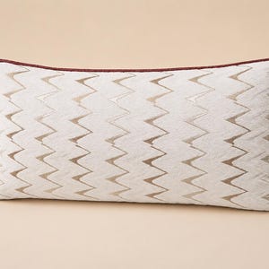 May include: A rectangular throw pillow featuring a cream and light brown chevron pattern across the front surface. The pillow is finished with a dark maroon piping trim along all four edges, providing a distinct border to the textured fabric.