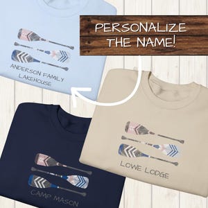 Peut inclure: Trois t-shirts, bleu clair, marine et beige, chacun avec un motif de trois rames aux détails aquarellés. Le t-shirt bleu clair dit "ANDERSON FAMILY LAKEHOUSE". Le t-shirt marine dit "CAMP MASON". Le t-shirt beige dit "LOWE LODGE".