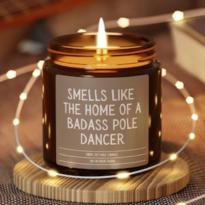 May include: A brown glass candle with a white label that reads "Smells like the home of a badass pole dancer". The candle is lit and sitting on a wooden coaster. The candle is surrounded by white string lights.