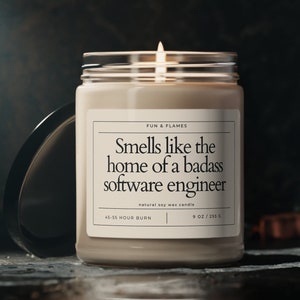 Puede incluir: Una vela de cera de soja beige en un tarro de vidrio con una tapa negra. La etiqueta de la vela dice "Smells like the home of a badass software engineer". La etiqueta también incluye "FUN & FLAMES", "natural soy wax candle", "45-55 HOUR BURN", y "9 OZ / 255 G".