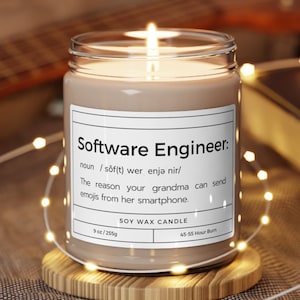 May include: A brown soy wax candle with a white label that reads "Software Engineer: noun /sof(t) wer enjə nir/ The reason your grandma can send emojis from her smartphone. SOY WAX CANDLE 9 oz/255g 45-55 Hour Burn"
