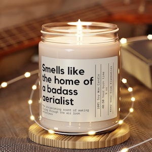 Puede incluir: Un tarro de vela de vidrio transparente con una vela de cera de soja rosa claro en su interior. La vela está etiquetada con "Smells like the home of a badass aerialist". La etiqueta también incluye detalles sobre la vela, como el tiempo de combustión y el hecho de que está hecha con 100% cera de soja. La vela está sentada sobre un posavasos de madera.