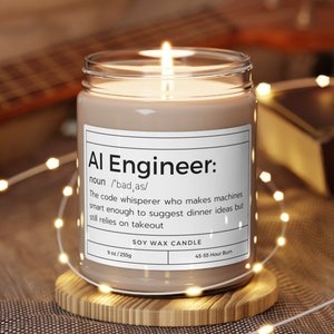 May include: A brown soy wax candle with a white label that reads "Al Engineer: noun /'bad as/ The code whisperer who makes machines smart enough to suggest dinner ideas but still relies on takeout SOY WAX CANDLE 9 oz/255g 45-55 Hour Burn".