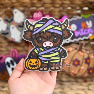 May include: A layered acrylic ornament featuring a brown Highland cow dressed as a mummy in purple and lime green glitter bandages, holding an orange jack-o'-lantern bucket. The text Desert Diva Co is printed in the lower right corner.