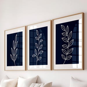 May include: Three framed prints with white line drawings of branches with leaves on a dark blue background. The prints are hanging on a white wall above a wooden bench with white cushions.