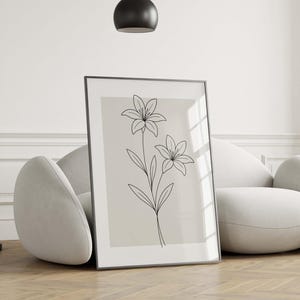 May include: A framed art print featuring a minimalist line drawing of two lilies. The black line art is set against a neutral background, encased in a black frame. The artwork is leaning against a modern, curved sofa.