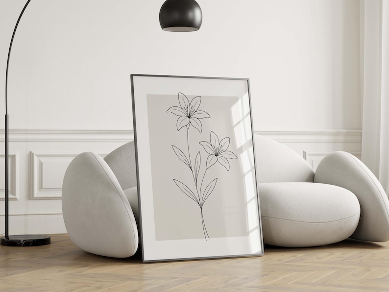 May include: A framed art print featuring a minimalist line drawing of two lilies. The black line art is set against a neutral background, encased in a black frame. The artwork is leaning against a modern, curved sofa.