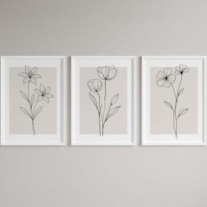 May include: Three framed floral art prints. Each print features a minimalist black line drawing of a different flower on a beige background. The frames are white, and the overall aesthetic is modern and elegant.