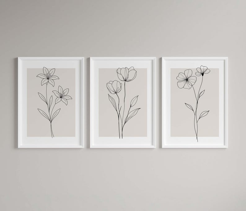 May include: Three framed floral art prints. Each print features a minimalist black line drawing of a different flower on a beige background. The frames are white, and the overall aesthetic is modern and elegant.
