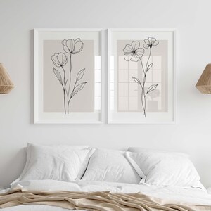 May include: Two framed floral art prints. Each print features a delicate black line drawing of flowers on a neutral background. The artwork is displayed above a bed with white linens, creating a serene and minimalist aesthetic.