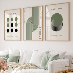 Könnte beinhalten: Drei gerahmte Bauhaus-Kunstdrucke mit geometrischen Designs in Schwarz und Olivgrün auf cremefarbenem Hintergrund. Die Drucke werden über einem weißen Sofa mit grünen und weißen Kissen präsentiert. Der Text "Bauhaus" und "Ausstellung 1923" ist sichtbar.