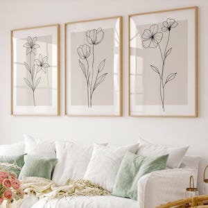 May include: Three framed botanical art prints. Each print features a delicate black line drawing of a different flower on a neutral background. The frames are a light wood colour, and the artwork is displayed above a white sofa with green and white cushions.