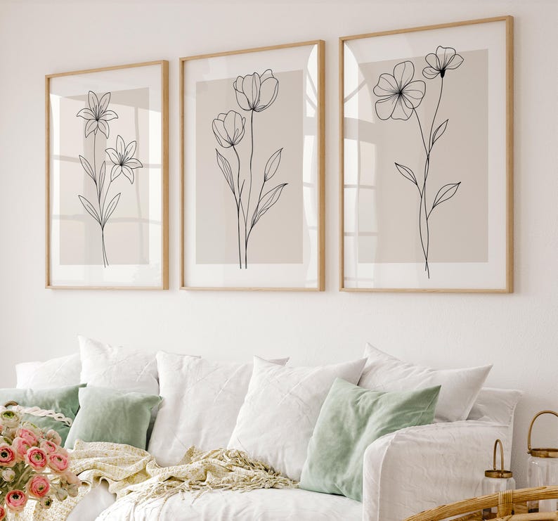 May include: Three framed botanical art prints. Each print features a delicate black line drawing of a different flower on a neutral background. The frames are a light wood color, and the artwork is displayed above a white sofa with green and white pillows.