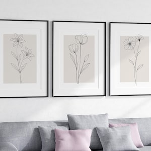 May include: Three framed floral art prints. Each print features a minimalist black line drawing of a different flower on a beige background. The frames are black, and the art is displayed above a gray sofa with pink and gray pillows.