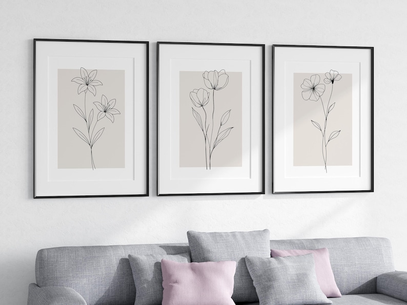 May include: Three framed floral art prints. Each print features a minimalist black line drawing of a different flower on a beige background. The frames are black, and the art is displayed above a gray sofa with pink and gray pillows.
