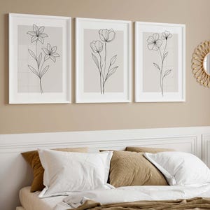 May include: Three framed botanical art prints with minimalist line drawings of flowers in white frames. The prints hang above a bed with white bedding and brown pillows. A round mirror and a lamp are also visible.