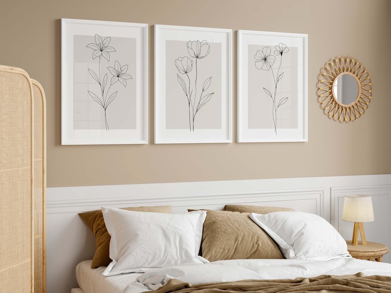 May include: Three framed botanical art prints with minimalist line drawings of flowers in white frames. The prints hang above a bed with white bedding and brown pillows. A round mirror and a lamp are also visible.