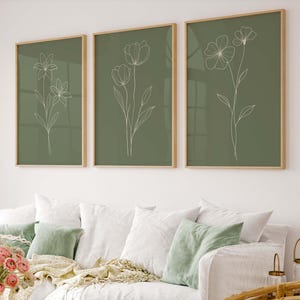 May include: Three framed botanical art prints. Each print features a white line drawing of a flower on a muted green background. The frames are a light wood color. The prints are displayed above a white couch with green pillows.