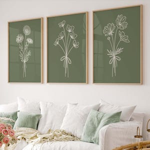 May include: Three framed botanical art prints. Each print features a white line drawing of a flower bouquet on a green background. The frames are a light wood color. The prints are displayed above a white couch with green pillows.