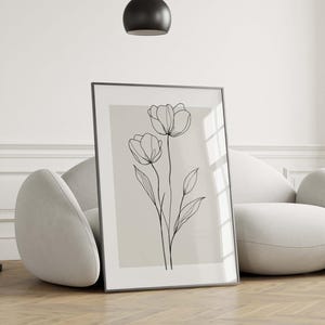 May include: A framed art print featuring a minimalist line drawing of two tulip flowers with stems and leaves. The artwork is in black against a beige background, set in a black frame. The print is leaning against a white curved sofa.