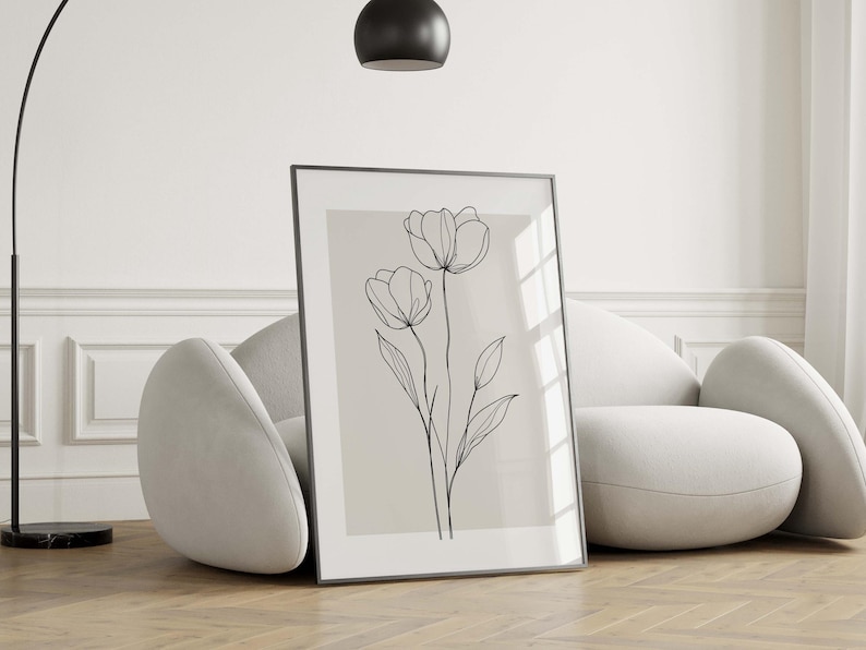 May include: A framed art print featuring a minimalist line drawing of two tulip flowers with stems and leaves. The artwork is in black against a beige background, set in a black frame. The print is leaning against a white curved sofa.