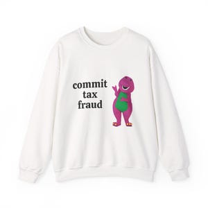 May include: A white sweatshirt featuring the phrase "commit tax fraud" and a cartoon dinosaur. The dinosaur is purple with a green belly and sandals. The sweatshirt is a casual, long-sleeved garment.