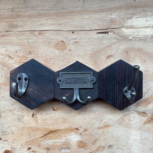May include: A rustic, wooden bathroom hook rack with a dark finish. The rack features three hexagonal panels joined together. Each panel has a metal hook, with the center panel displaying the word "BATHROOM".