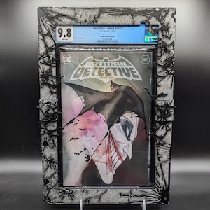 Puede incluir: Un cómic calificado en una caja protectora. La portada del cómic presenta una ilustración de acuarela de Batman y el Joker. El cómic se titula "Batman Detective Comics" y el número es 1027. El cómic está calificado con 9.8.