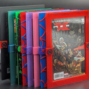 Puede incluir: Una colección de marcos para cómics en varios colores, incluyendo rojo, verde y azul. Cada marco presenta un diseño único y un cierre con un logotipo de superhéroe. Los marcos contienen cómics, uno de ellos muestra la portada de "Ultimate X-Men" número 75.
