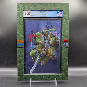 Puede incluir: Una placa de cómic verde clasificada con una calificación de 9,8. La portada del cómic presenta Teenage Mutant Ninja Turtles #1, con las cuatro tortugas en acción. La placa tiene un patrón de caparazón de tortuga verde y negro.