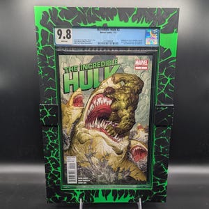 Puede incluir: Un protector de cómic verde y negro con un cómic clasificado en su interior. El cómic es "The Incredible Hulk #2" y tiene una calificación de 9,8. La portada del cómic muestra a Hulk luchando contra un tiburón gigante.