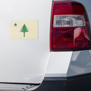 May include: A rectangular car magnet displaying the flag of Maine. The design features a green pine tree and a blue star on a cream background. The magnet is attached to the white bodywork of a vehicle.